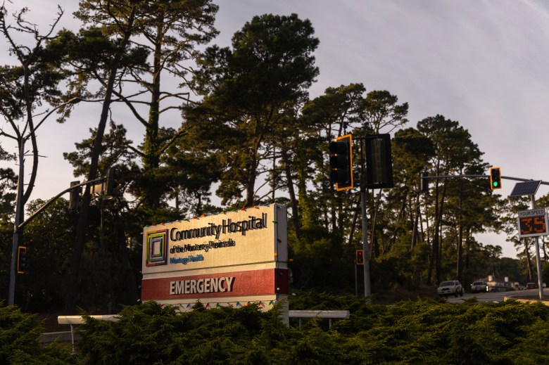 A sign that reads "Community Hospital of the Monterey Pennisula" is visible in between green foliage and in front of trees in Monterey. The road has stoplights that lead to the hospital.