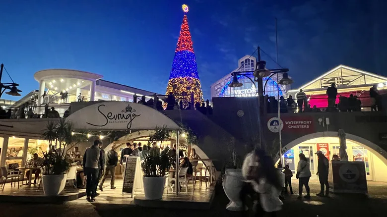 A Christmas tree decorated with gold, red, and blue lights in a shopping mall in Cape Town, South Africa