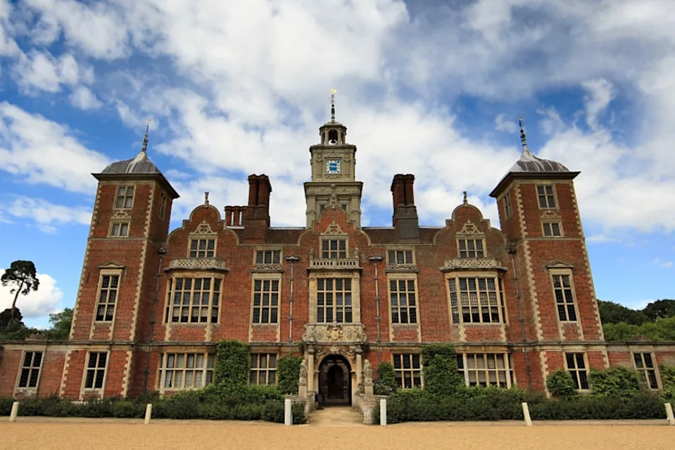 A large, historic brick mansion with multiple chimneys and a clock tower, set against a partly cloudy sky