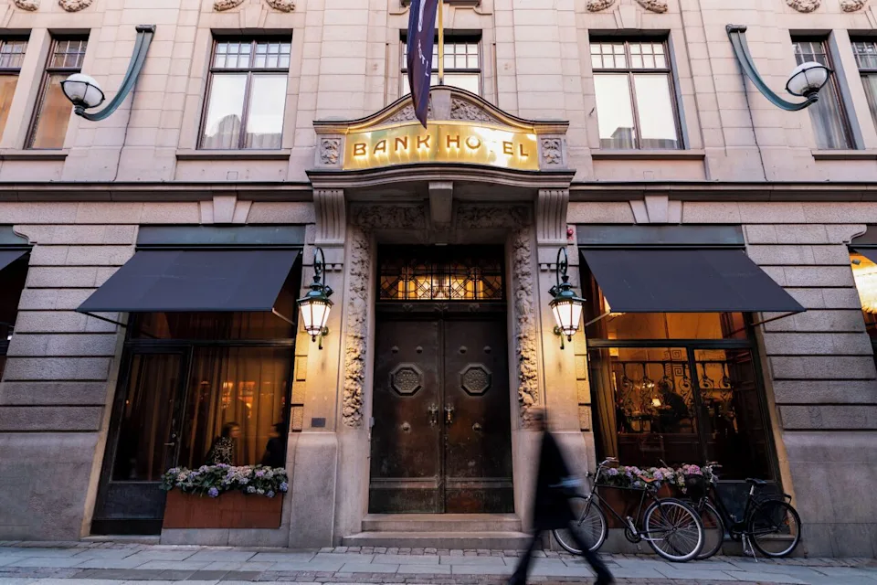 View of the entrance to the Bank Hotel with blurred person walking by and bicycles parked in front, the two imposing metal doors are apparent that it was historically a bank building. - Courtesy of the Bank Hotel