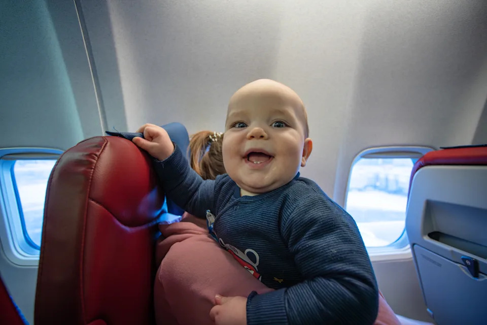 A joyful baby sits on a parent's lap aboard an airplane, smiling and enjoying the flight. The cozy cabin and window light create a warm, family travel moment.