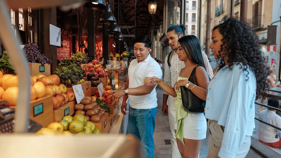 Group of friends enjoying their vacation in Spain, selecting vibrant exotic fruits at a lively market stall filled with fresh produce