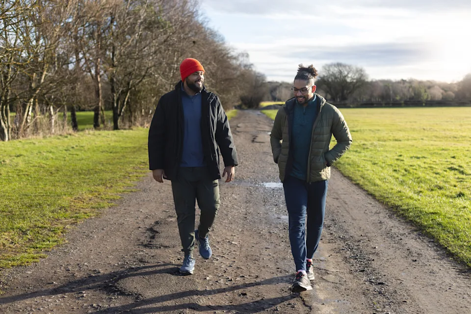 A front full-length view of two mid-adult multiracial male friends walking side by side along a dirt track on a cool day in Gosforth, England. They are wearing casual, warm outdoor clothing. The natural setting and open space convey calm, reflection, and connection. The image represents friendship, open conversation, mental health awareness, and the importance of staying active for overall well-being.Videos are available similar to this scenario.