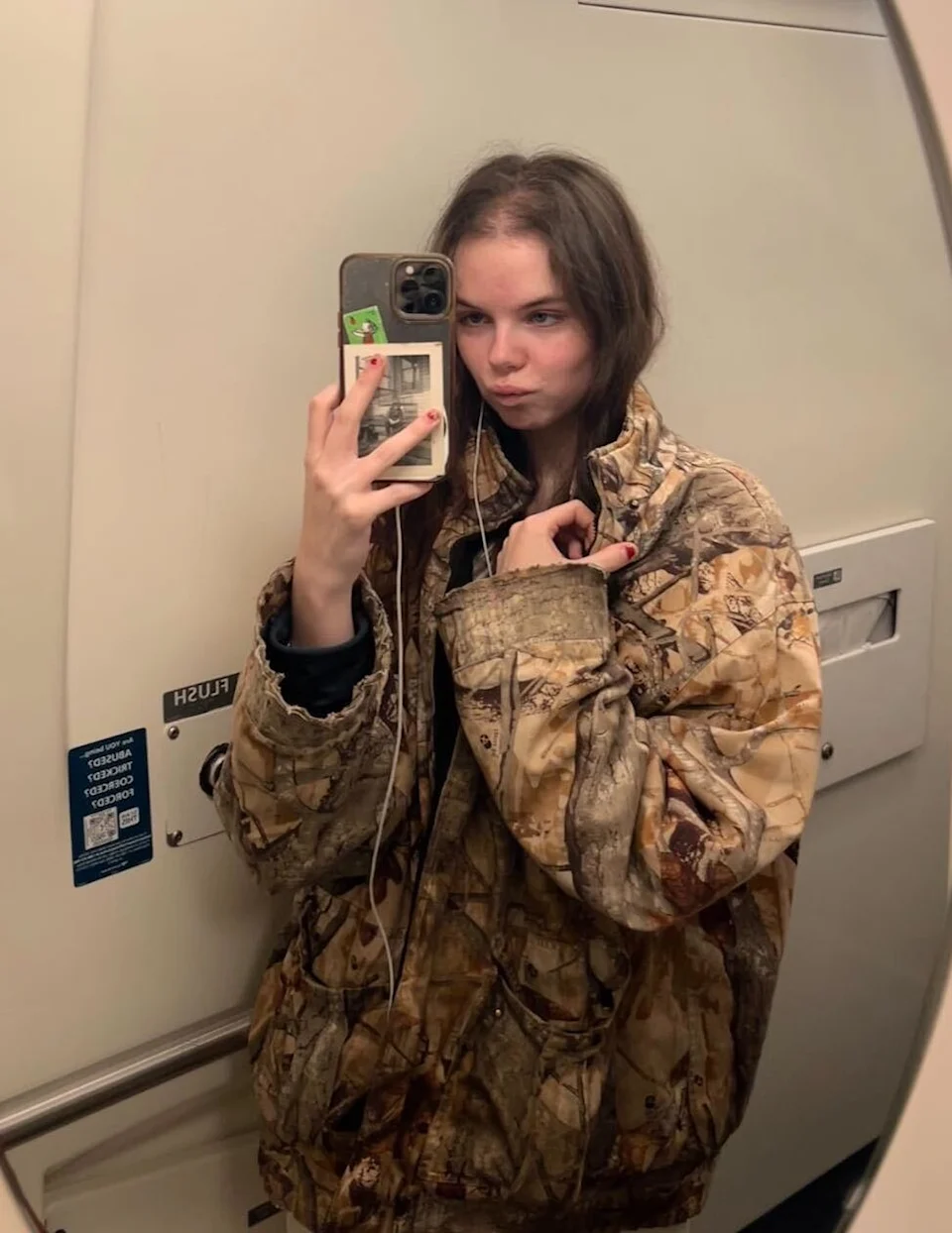 Person taking a mirror selfie in a train bathroom, wearing a large camouflage jacket and holding a smartphone.
