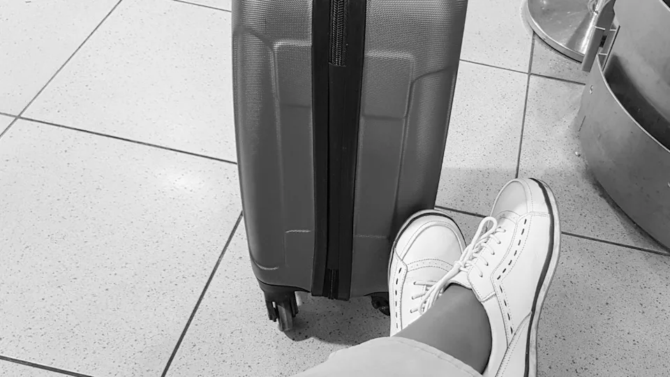 A black-and-white photo of a woman's leg in trousers and sneakers, sitting in a relaxed pose, with her legs placed near a suitcase, waiting for a trip.