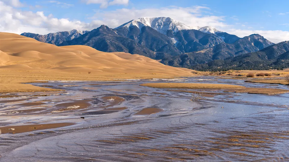A bright sunny Spring morning view of Medano Creek rushing down a sandy valley at base of rolling Great Sand Dunes and snow-capped Mt. Herard. Great Sand Dunes National Park, Colorado, USA.