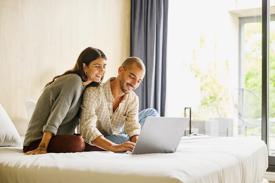 Medium wide shot of smiling couple relaxing on bed using laptop to plan vacation travel