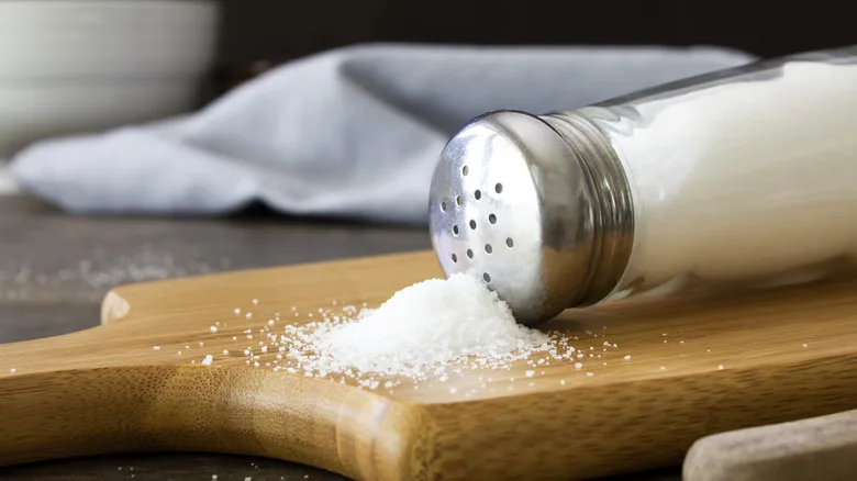 A shaker of table salt spilled on a cutting board