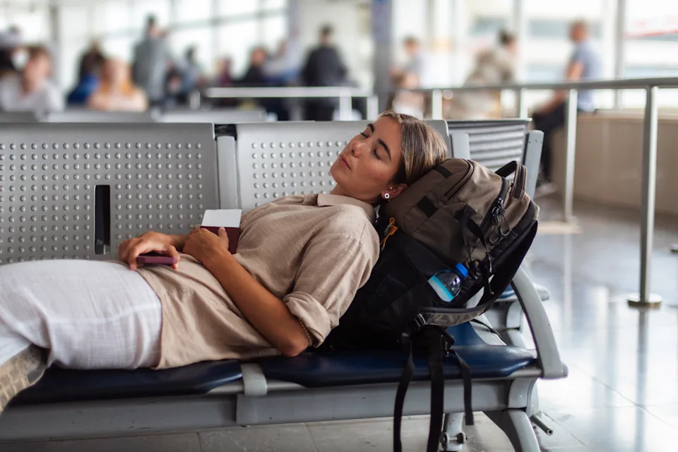 A weary traveler rests on a bench at a busy airport, backpack and passport at hand; Calm moment amid bustle, capturing travel fatigue, relaxation, and everyday journey
