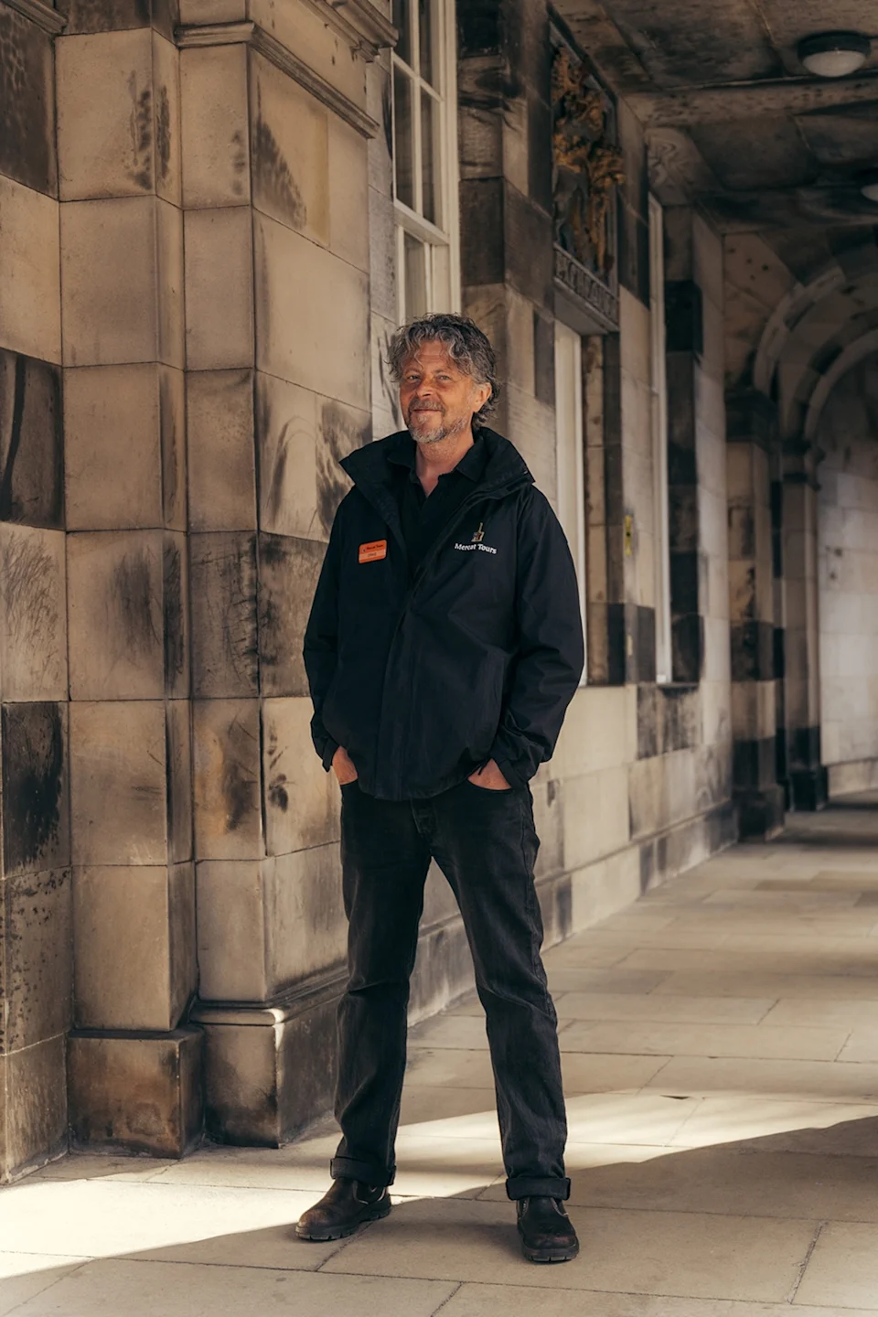 A friendly local, middle-aged man in dark weather-proof attire standing in an old stone corridor.