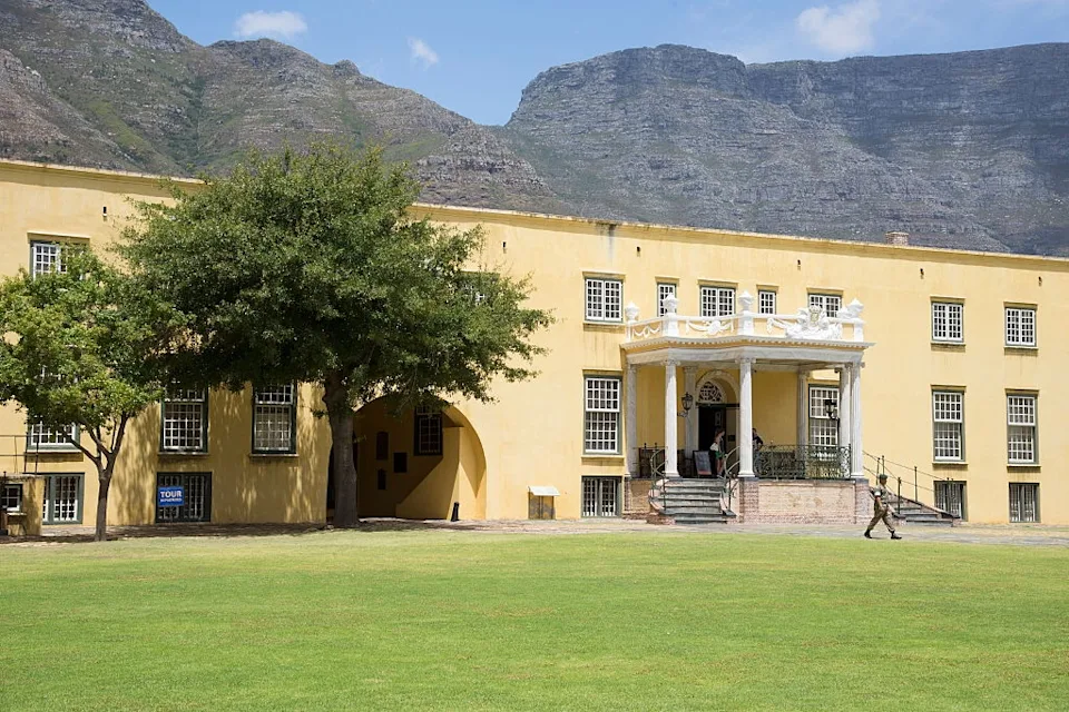 Historic yellow building with arched windows and a grand entrance, set against a mountainous backdrop. A person walks on the lawn