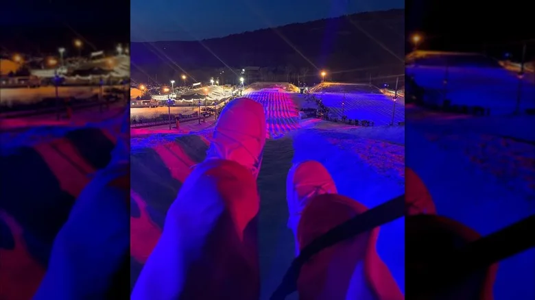 Nighttime view from a lift at Blue Mountain Resort, with legs in the foreground and colorful lights on the snow tubing lanes below