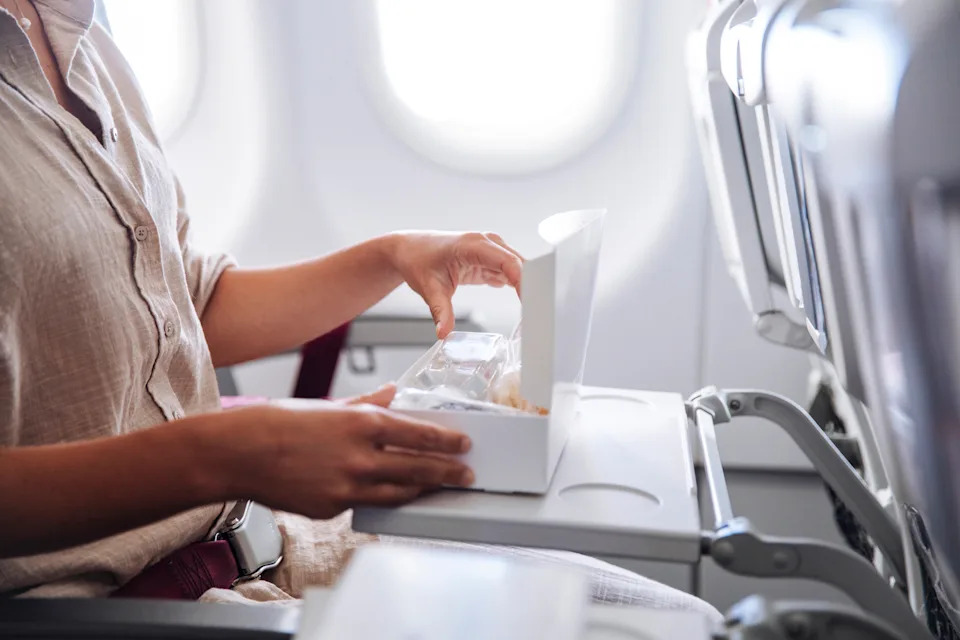 A traveler seated on an airplane opens a small meal box from an in-flight lunch; The relaxed, everyday moment captures travel, dining, and a sense of solitude at cruising altitude