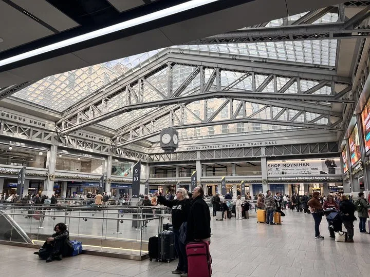 Interior of Moynihan Train Hall in New York City, showing travelers with luggage beneath a large glass ceiling and metal beams, with station signs and shops visible in the background.