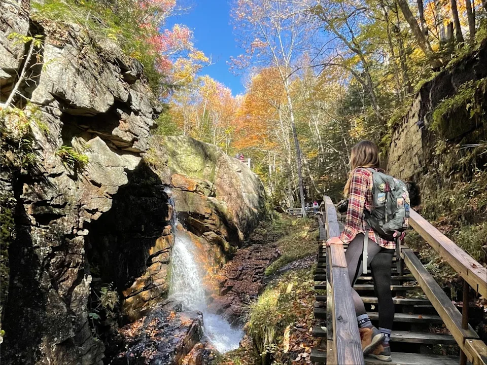 Emily climbs up a flight of wooden stairs while looking over at a waterfall and fall foliage.