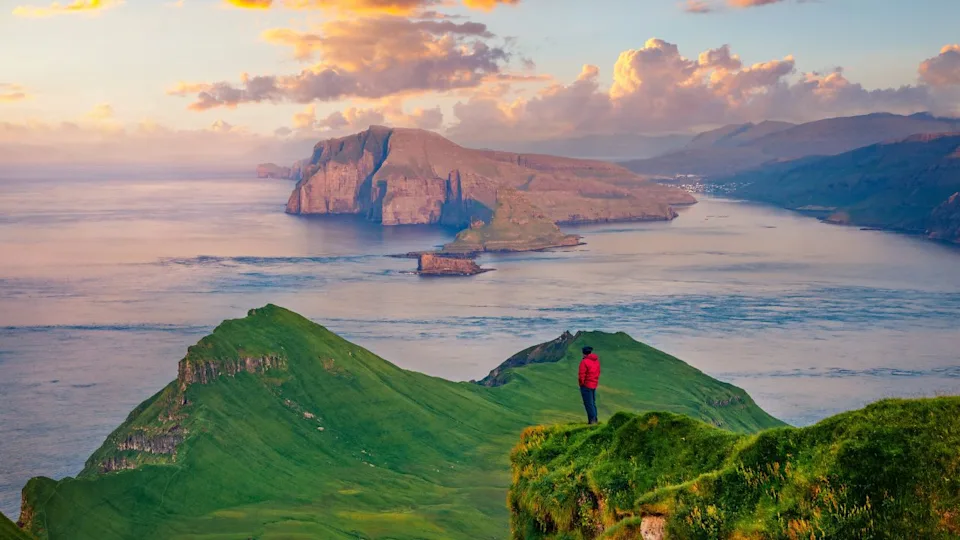 Tourist in red jacket admiring of sunset on Alaberg cliffs, Faroe Islands, Denmark, Europe. Aerial morning view of Mykines island with Vagar island on background. Great Atlantic seascape.
