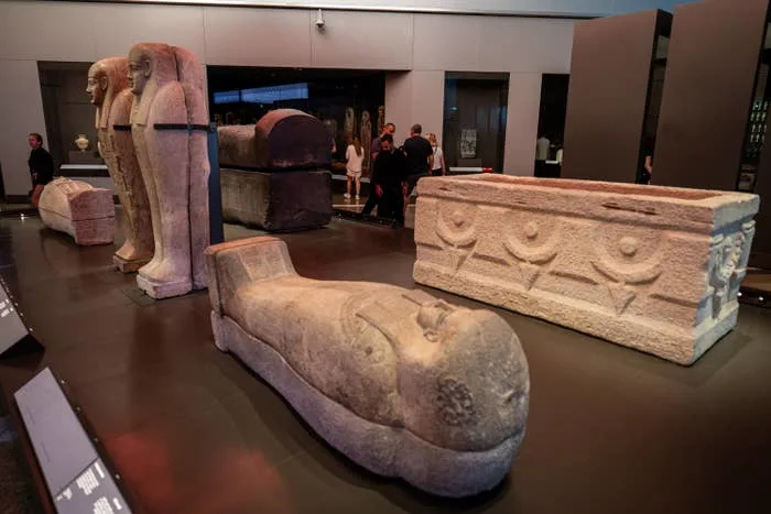 Ancient Egyptian sarcophagi and artifacts displayed in a modern museum exhibit with visitors in the background