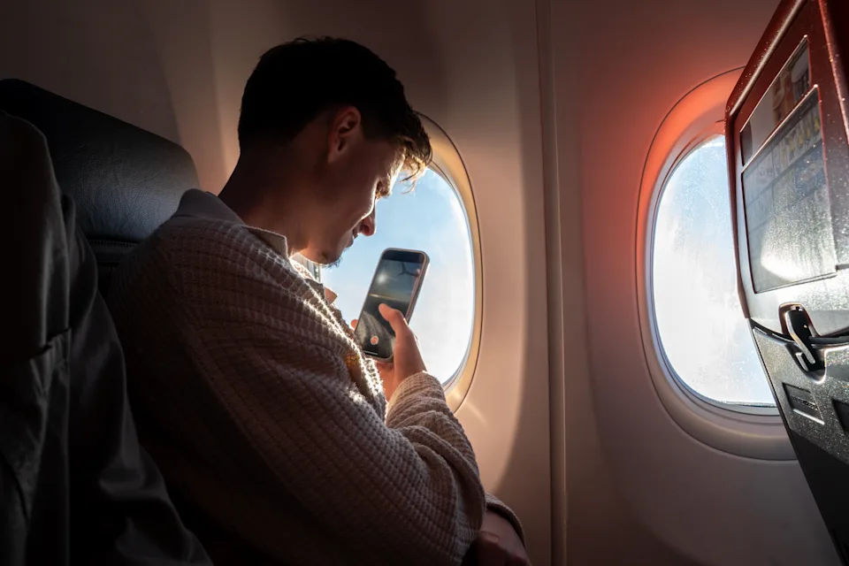 A waist-up shot of a male passenger taking a picture on their phone out of an aeroplane window. He is wearing comfortable casual clothing.