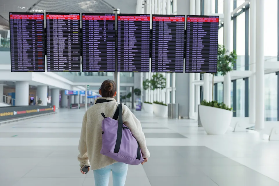 A traveler moves through a modern airport terminal, focusing on electronic departure boards displaying flight information. The spacious area features large windows and greenery.
