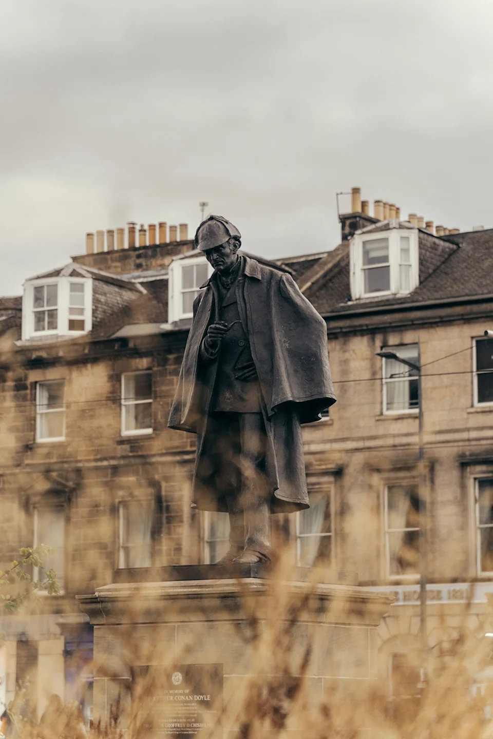 A bronze statue of Detective Sherlock Holmes in a short cape, shot through dry, tall grass with Edinburgh's stone house in the background.