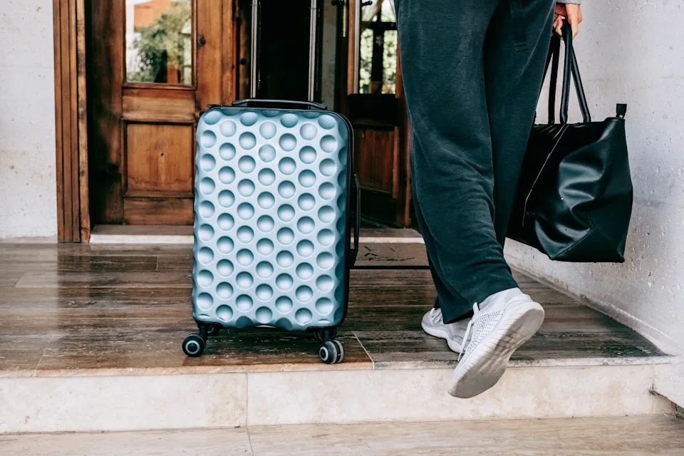 Woman wheels a blue suitcase and carries a black leather duffel in a door. Check-in, onward travel, weekend vacation concept.