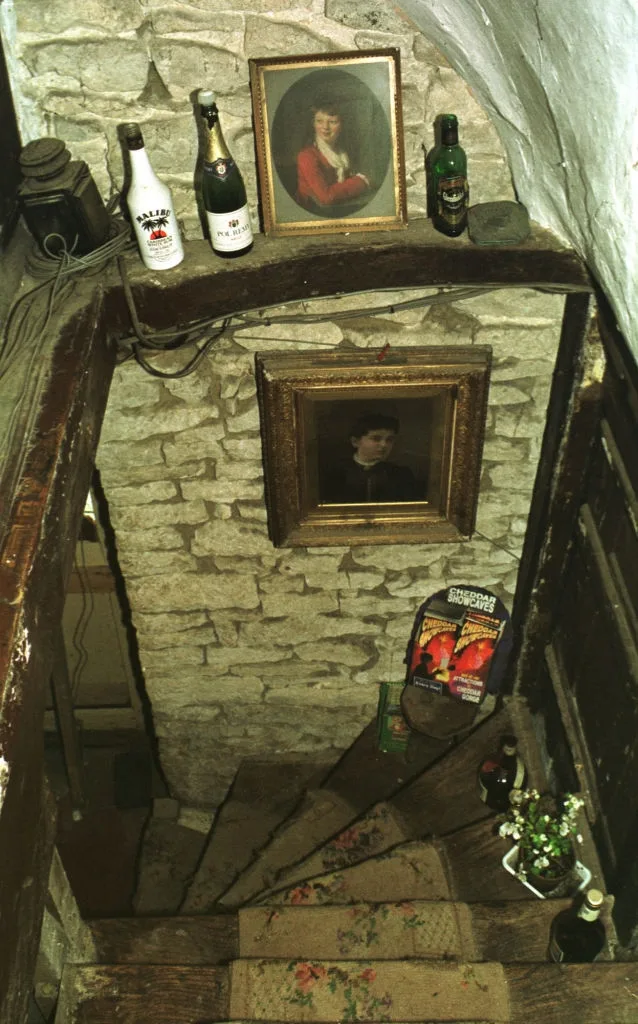 Stone staircase with vintage portraits, bottles, and a poster on the walls. Flowers in a vase are placed at the base of the stairs