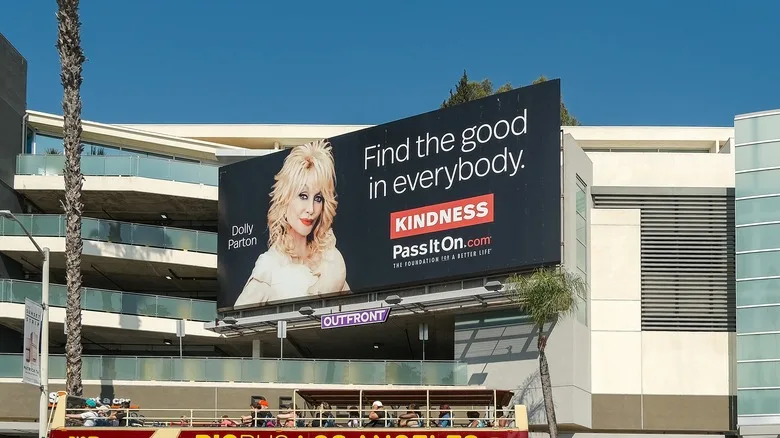 A billboard featuring Dolly Parton saying "find the good in everybody" above a busy LA street