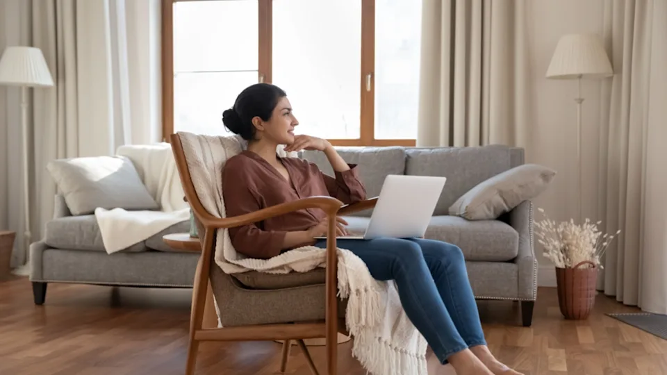 Smiling dreamy Indian woman using laptop, sitting in armchair at home, looking in distance, happy thoughtful young female freelancer looking in distance