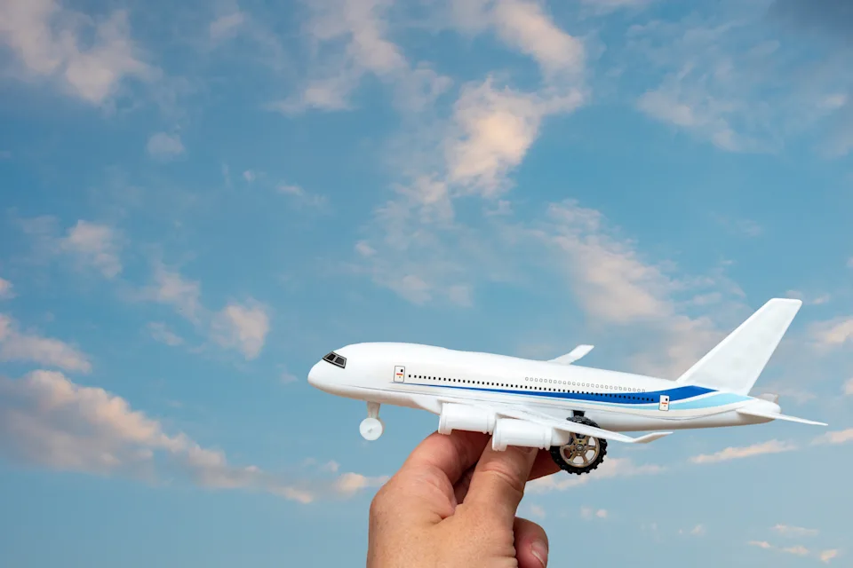 A hand holding a toy white airplane against the backdrop of a beautiful sky with clouds illuminated by the setting sun, copy space. Travel, vacation concept.