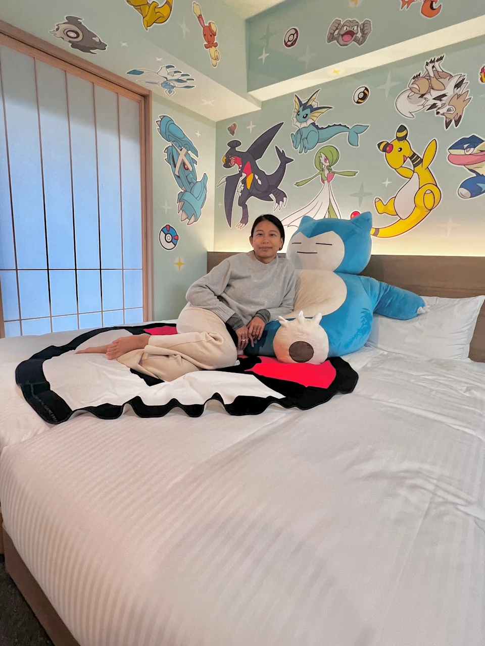 Person sitting on a bed with a large plush toy in a room decorated with animated character art on the walls