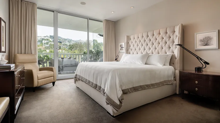 The bedroom features a large bed with a soft headboard and a sliding glass door.