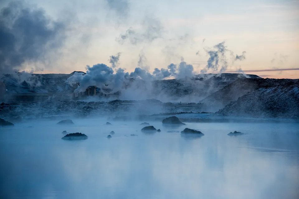 Steam rises from a geothermal hot spring surrounded by rocky terrain, with a hazy sky above, creating a tranquil and mysterious atmosphere