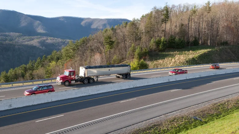 A highway with a semitruck and cars next to rolling hills and trees.