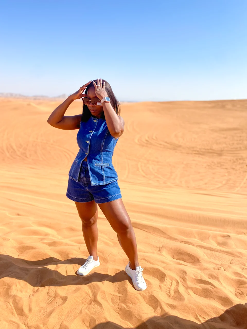 I stand on the Dubai desert sand wearing a sleeveless denim romper, white sneakers, and sunglasses under the clear blue sky.