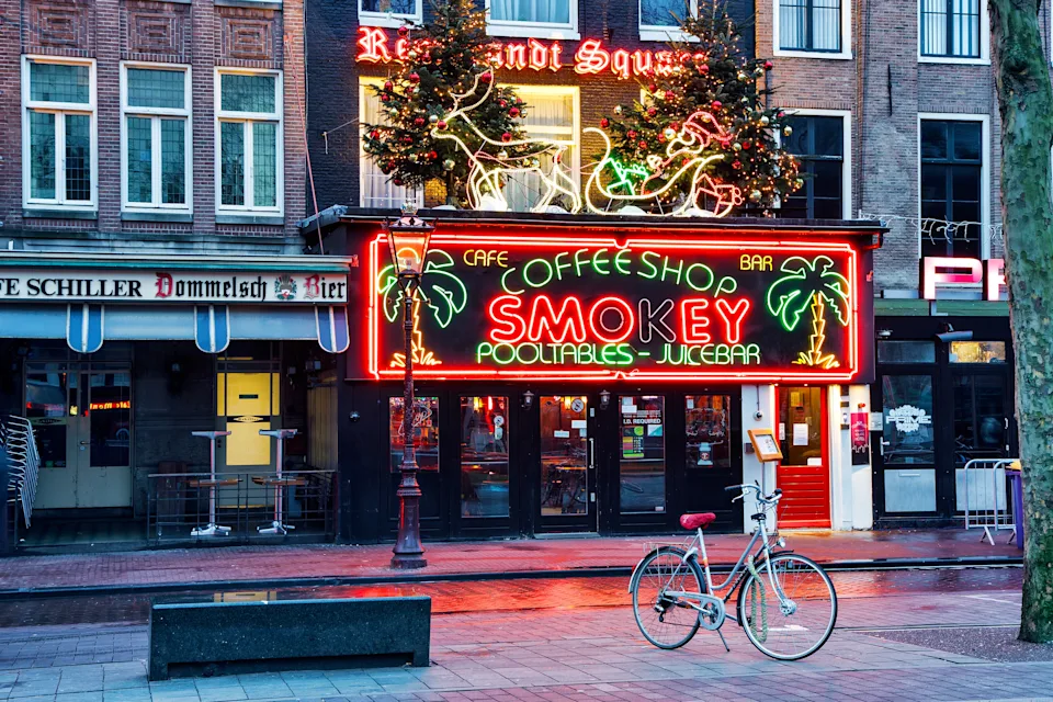 A lively street scene in Amsterdam featuring a neon-lit coffee shop sign with palm trees, bicycles parked outside, and festive lights above