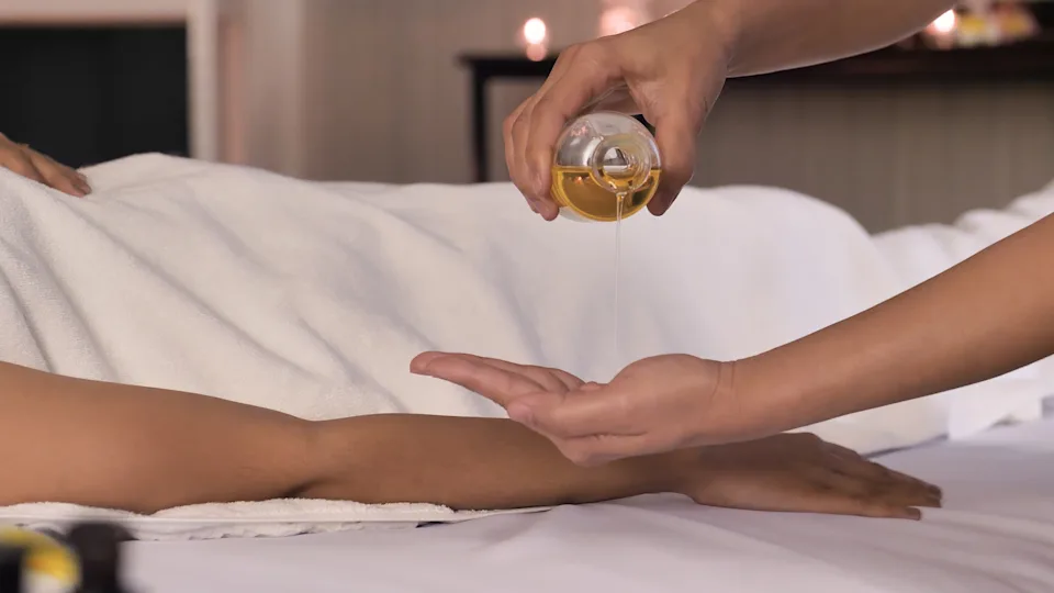Person pouring oil into their hand, preparing for a massage on a person lying on a table covered with a towel, implying relaxation and self-care