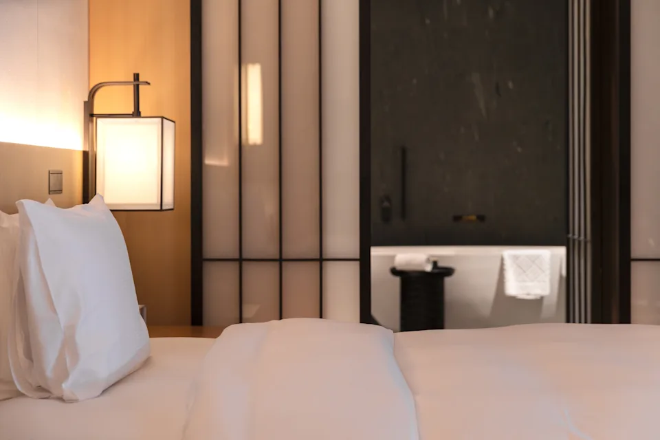 Elegant hotel room with a neatly made bed, modern lamp, and view into a bathroom featuring a towel hanging by a bathtub