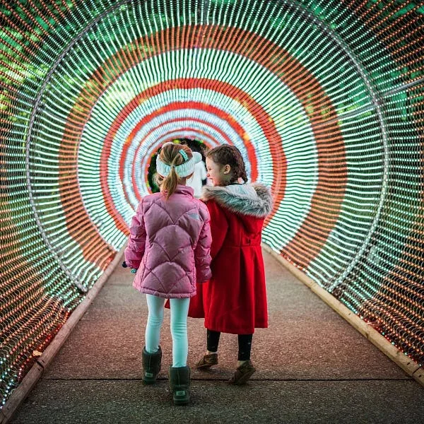 two children in winter clothing standing in a colorful illuminated tunnel illuminated by red, green and white striped lights