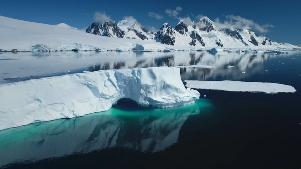 Huge towering iceberg in Antarctica floats polar ocean in sunny day. Snow covered mountain range and glacier in background reflect in crystal water. Arctic winter landscape at global warming problem.