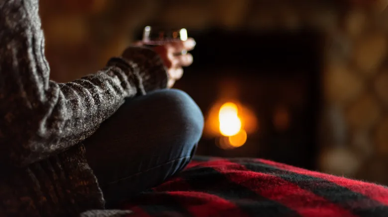 Close up of a glass of wine in hand, cozy blanket, and a burning fireplace