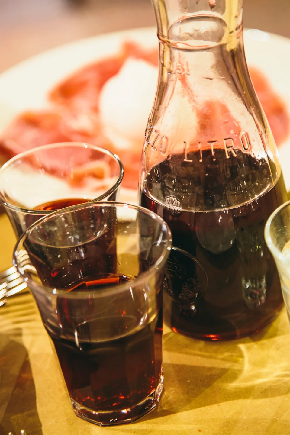 Carafe and glasses of red wine on a table with a plate of sliced meats in the background, highlighting a dining experience
