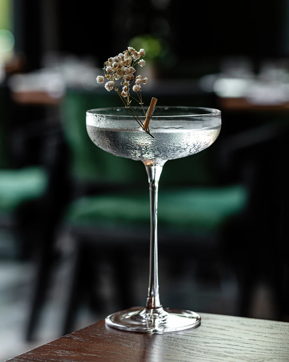 A sophisticated cocktail in a coupe glass, garnished with delicate baby's breath, sits on a wooden table in a dimly-lit upscale setting