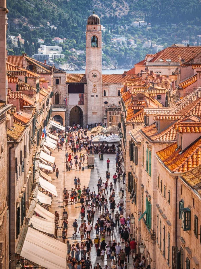 Aerial view of a bustling street in Dubrovnik, Croatia, with people walking and historic buildings lining the path. Tower with a clock in the background