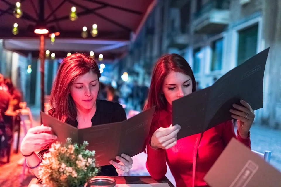 Two people sitting at an outdoor cafe, looking at menus under an umbrella, with a small plant on the table