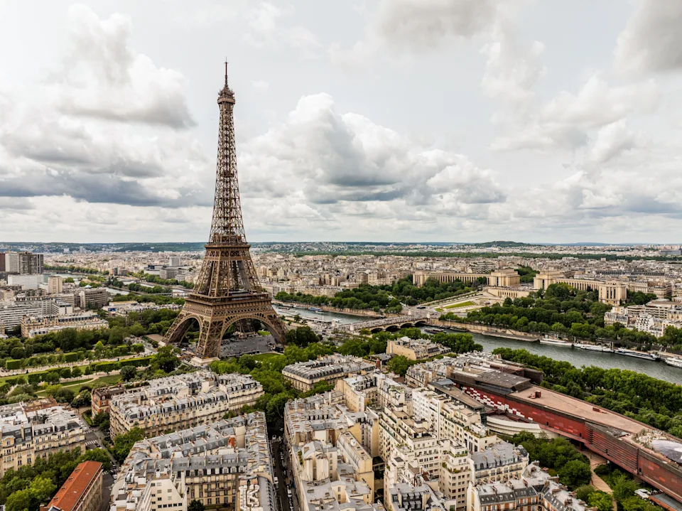 Aerial view of Paris with the Eiffel Tower in the center, surrounded by cityscape and the Seine River, under a partly cloudy sky