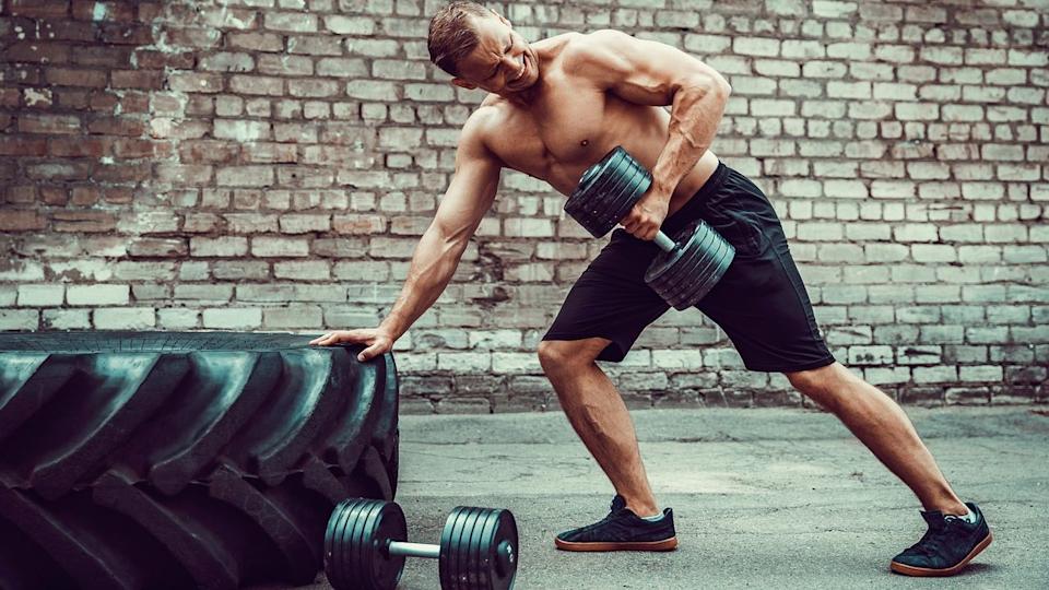 Man leaning against a tyre with one hand and performing a dumbbell bentover row with his left arm.