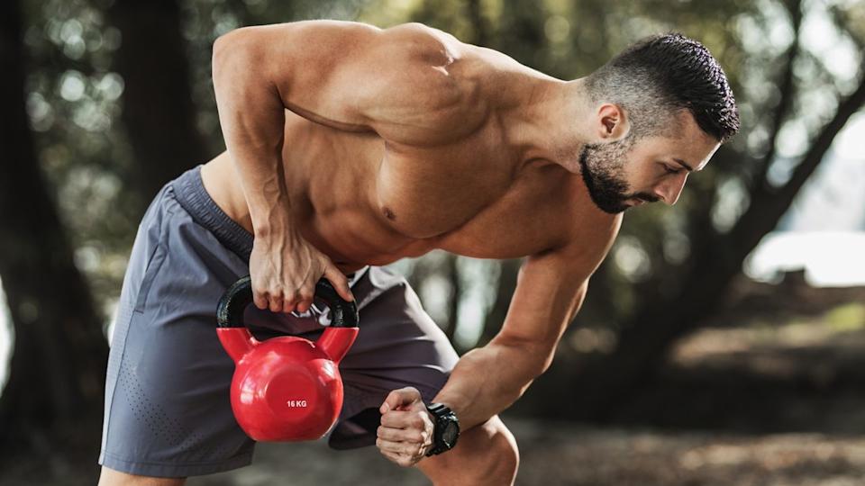 Muscled male rowing kettlebell with right arm outdoors.