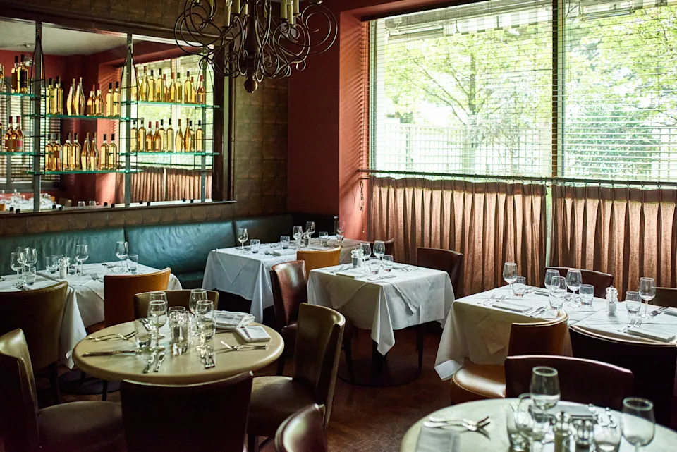 Chic restaurant interior with elegantly set tables, wine bottles on shelves, and a large window letting in natural light, enhancing a cozy ambiance