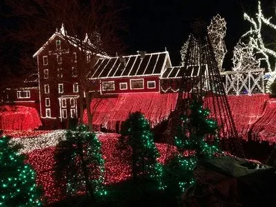 mill buildings outlined in white christmas lights with trees in white lights beside and behind it and green lit trees in the foreground, there are thousands of red christmas lights pouring down in a waterfall effect from the mill