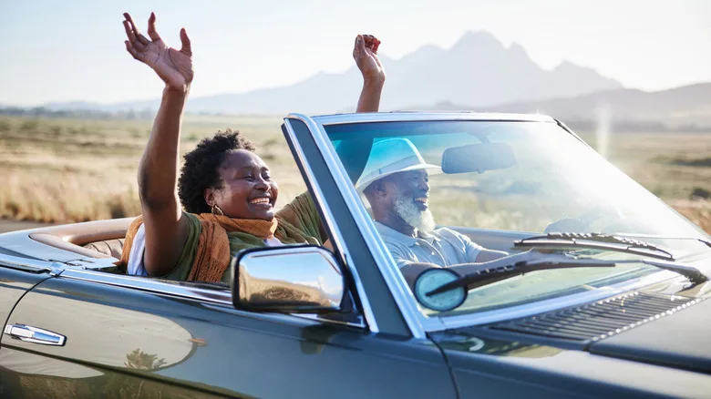 A happy retired couple driving a convertible with the top down where the man is driving and the woman has her hands raised in the air.